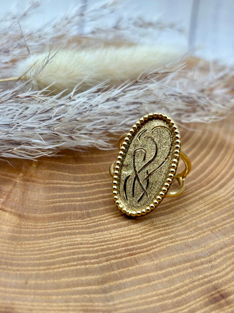K&ouml;nnte beinhalten: Goldfarbener Ring mit ovaler Oberfl&auml;che und gepr&auml;gtem Design. Der Ring hat einen Perlenrand und ein dekoratives Band. Der Ring wird auf einer Holzoberfl&auml;che pr&auml;sentiert.
