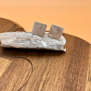May include: A pair of silver square earrings with a textured finish. The earrings are displayed on a piece of driftwood.