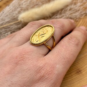K&ouml;nnte beinhalten: Ein goldfarbener Ring mit ovalem Gesicht, das ein strukturiertes Design und einen Perlenrand aufweist. Der Ring hat einen doppelb&auml;ndigen Schaft. Der Ring befindet sich an einer Hand.