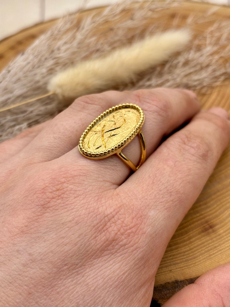 K&ouml;nnte beinhalten: Ein goldfarbener Ring mit ovalem Gesicht, das ein strukturiertes Design und einen Perlenrand aufweist. Der Ring hat einen doppelb&auml;ndigen Schaft. Der Ring befindet sich an einer Hand.