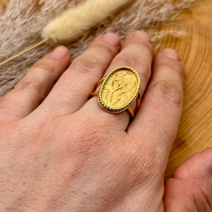 K&ouml;nnte beinhalten: Ein goldfarbener Ring mit einem ovalen Gesicht mit einem gepr&auml;gten Design. Der Ring hat ein strukturiertes Band und einen detaillierten Rand. Der Ring befindet sich an einem Finger. Das Design des Rings ist Vintage-Stil.