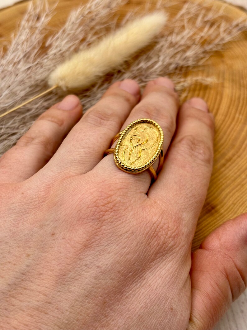 K&ouml;nnte beinhalten: Ein goldfarbener Ring mit einem ovalen Gesicht mit einem gepr&auml;gten Design. Der Ring hat ein strukturiertes Band und einen detaillierten Rand. Der Ring befindet sich an einem Finger. Das Design des Rings ist Vintage-Stil.