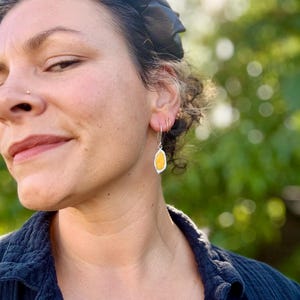 Op de afbeelding: Zilveren oorbellen met een gele, druppelvormige steen. De steen is gezet in een zilveren rand en hangt aan een zilveren ring. De achtergrond is een wazig beeld van groen gebladerte.