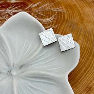 May include: A pair of silver square earrings with a textured, geometric design. The earrings are displayed on a white ceramic dish.