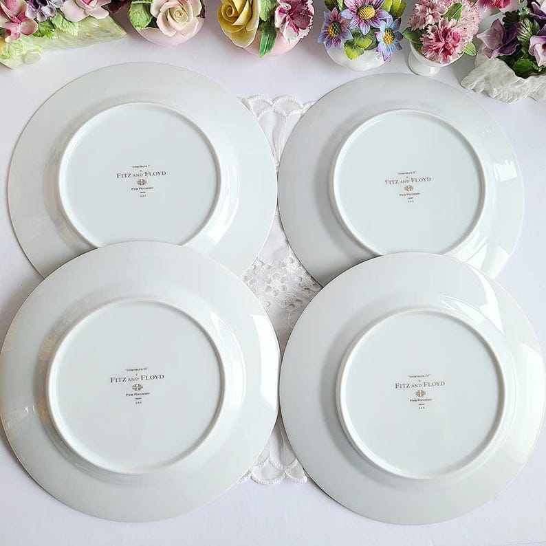 May include: Four white Fitz and Floyd dinner plates, arranged in a square, with the brand name and design details visible on the underside. The plates are on a white lace surface, with floral decorations in the background.