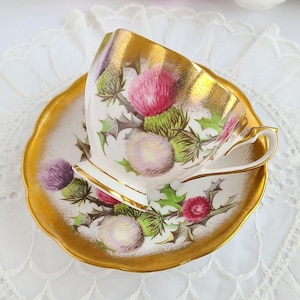 May include: A white teacup and saucer set with a gold rim and a floral thistle design in pink, purple, and green. The teacup has a gold interior and handle. The saucer has a scalloped edge.