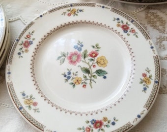 May include: A white ceramic plate with a floral design. The plate has a brown and green border with a delicate floral pattern. The center of the plate features a larger floral design with pink, yellow, and blue flowers.