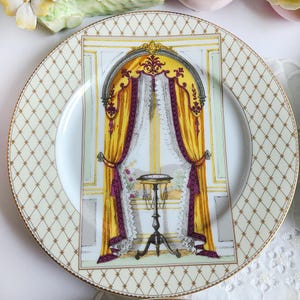 May include: A decorative porcelain plate with a gold rim and a cream-colored background featuring a repeating diamond pattern. The center of the plate showcases a detailed illustration of a window with yellow curtains, purple accents, and a small table.
