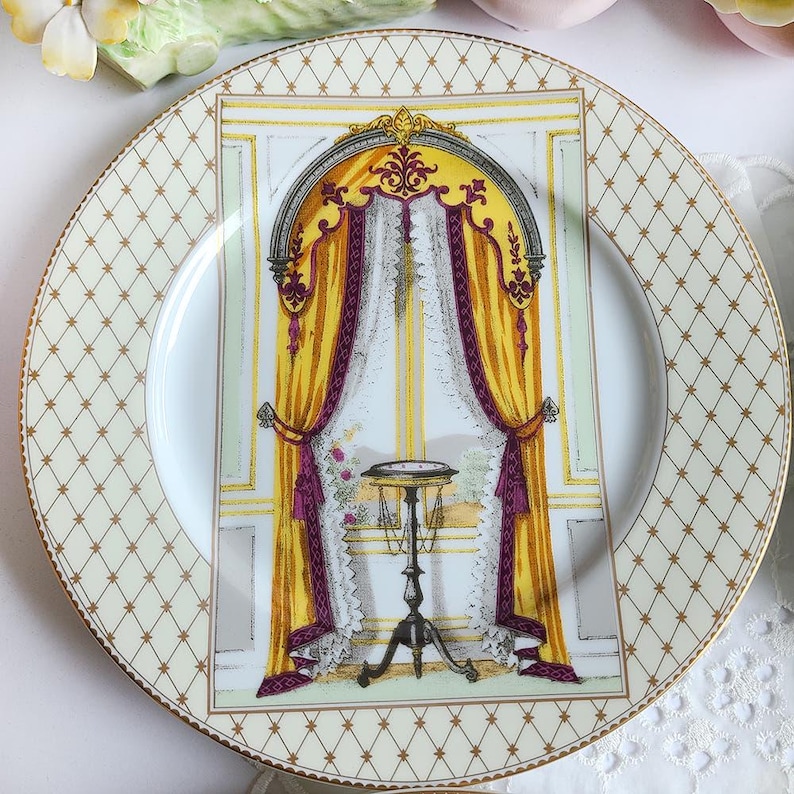 May include: A decorative porcelain plate with a gold rim and a cream-colored background featuring a repeating diamond pattern. The center of the plate showcases a detailed illustration of a window with yellow curtains, purple accents, and a small table.