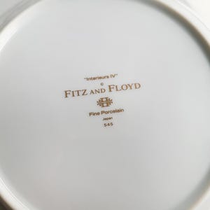 May include: White porcelain plate with gold text and a decorative emblem. The plate features the text "Interieurs IV", "FITZ AND FLOYD", "Fine Porcelain", "Japan", and "545".