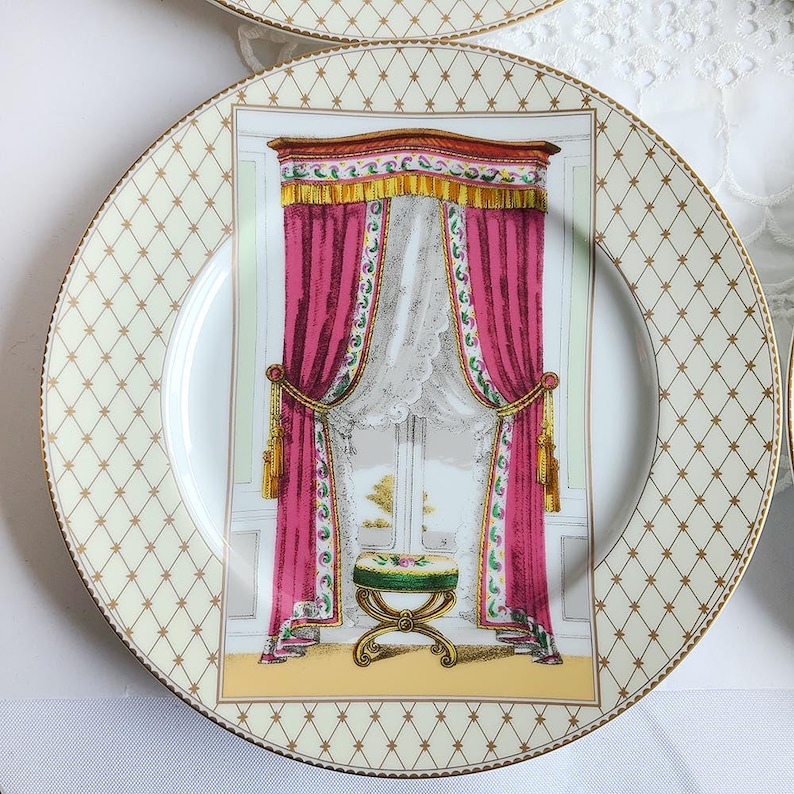 May include: A decorative plate with a cream-colored background and a gold diamond pattern. The center features a detailed illustration of a window with pink curtains, a white lace curtain, and a small stool.