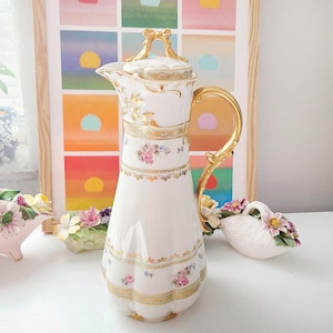 May include: A white porcelain coffee pot featuring a gold-toned handle and a lid topped with a gold bow-shaped finial. The body is decorated with delicate floral patterns in pink, blue, and green, accented by horizontal gold bands and fluted details.