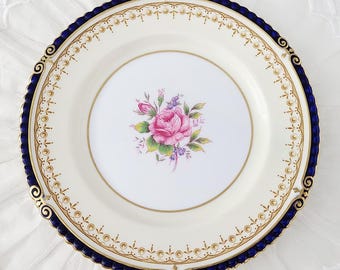 May include: A white porcelain plate with a pink rose floral design in the center. The plate has a navy blue rim with gold accents and a gold border. The plate is round and has a scalloped edge.