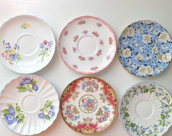 May include: Six round ceramic saucers arranged in two rows. The saucers feature various floral patterns, including blue daisies, pink roses, and blue morning glories, with some plates having scalloped edges and others featuring gold trim details.