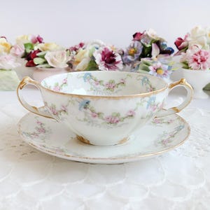 May include: A white porcelain teacup and saucer set with a floral design in pink, blue, and green. The teacup has two handles and gold trim. The saucer also has a matching floral pattern and gold trim. The set is on a white lace tablecloth.