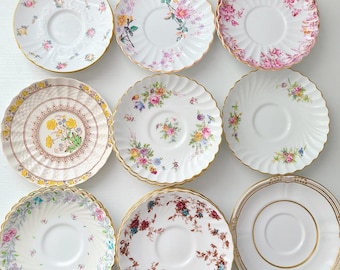 May include: A collection of nine round ceramic saucers arranged in a grid. Each saucer features a unique design, including various floral patterns in pink, yellow, and green, scalloped edges, and gold-toned rims on several pieces.