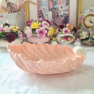 May include: A light peach-colored, ceramic serving dish with a shell-like design. The dish has a scalloped edge and a decorative, white, spiral accent. The background includes floral arrangements and a vintage clock.