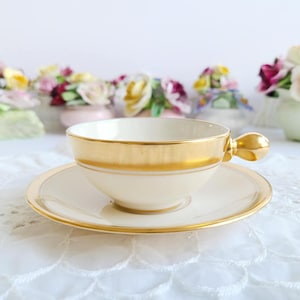 May include: Cream-colored teacup and saucer set with gold trim. The teacup has a rounded shape and a gold handle. The saucer is round and has a matching gold trim. The set is placed on a white surface.