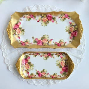 May include: Two rectangular serving trays with gold rims and handles. The trays are white with a floral design of pink and peach roses. The trays are placed on a white, lacy surface.