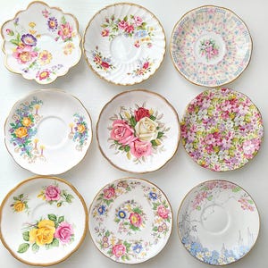 May include: A collection of nine ceramic saucers arranged in a grid, each featuring distinct floral patterns in various colors including pink, yellow, blue, and green, with gold-toned rims on several pieces and scalloped edges.