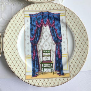 May include: A decorative plate with a cream-colored background and a gold geometric pattern around the rim. The center features a detailed illustration of a room with blue curtains, a green chair, and a patterned wallpaper.