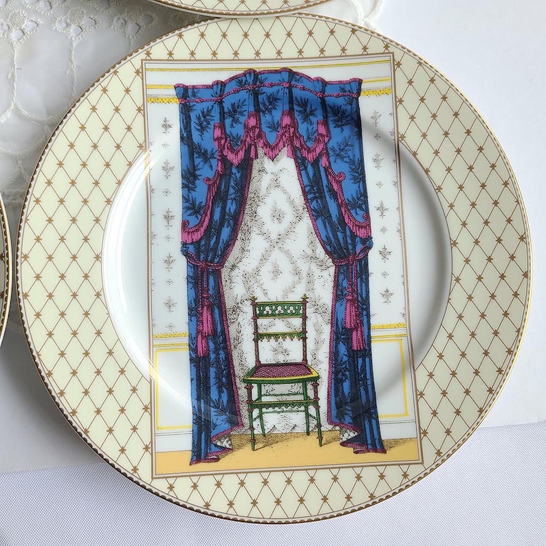 May include: A decorative plate with a cream-colored background and a gold geometric pattern around the rim. The center features a detailed illustration of a room with blue curtains, a green chair, and a patterned wallpaper.