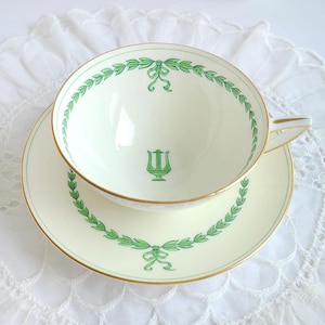 May include: Cream-colored teacup and saucer set with green leaf and bow design. The cup and saucer have gold trim. A green lyre symbol is in the center of the cup. The set is on a white lace surface.