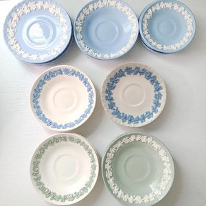 May include: A collection of ceramic saucers featuring raised white floral relief patterns along the rims. The set includes light blue, cream, and pale green saucers, each with a central circular indentation for a cup, showcasing classic stoneware.