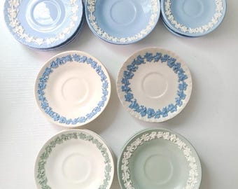 May include: A collection of ceramic saucers featuring raised white floral relief patterns along the rims. The set includes light blue, cream, and pale green saucers, each with a central circular indentation for a cup, showcasing classic stoneware.