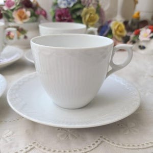 May include: A white teacup and saucer set. The teacup has a ribbed design and a curved handle. The saucer has a decorative, embossed pattern around the edge. The set is on a white tablecloth, with other teacups and floral arrangements in the background.