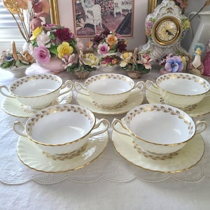May include: Six cream-colored teacups and saucers with gold leaf trim. The teacups have two handles and sit on matching saucers. The rims and handles are accented with gold. The set is arranged on a white surface.