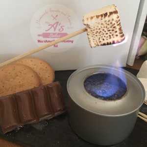 May include: A marshmallow roasting kit with a small, portable butane burner, two chocolate bars, and two biscuits. The marshmallow is on a skewer over the flame.
