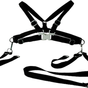 Puede incluir: Arnés negro con múltiples correas, anillos metálicos y una hebilla plateada. El arnés tiene correas para los hombros conectadas a una correa para el pecho. Correas adicionales se extienden hacia abajo con anillos metálicos.