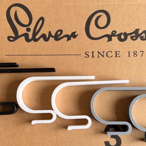 May include: Four metal stocking holders in black, white, and gray, arranged in a row. The holders are in a curved, hook-like shape. The Silver Cross logo and "SINCE 1877" are visible in the background.