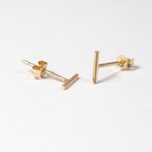 May include: A pair of gold-colored bar stud earrings. Each earring features a small, straight bar design. The earrings are displayed on a white background, highlighting their simple, minimalist style. These earrings are a classic jewelry accessory.