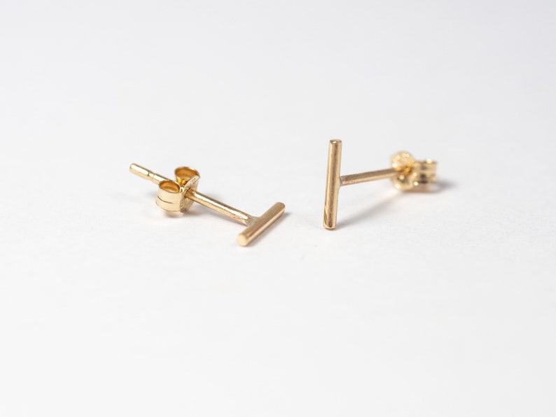 May include: A pair of gold-colored bar stud earrings. Each earring features a small, straight bar design. The earrings are displayed on a white background, highlighting their simple, minimalist style. These earrings are a classic jewelry accessory.