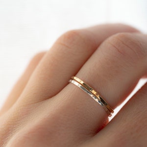 Puede incluir: Dos anillos finos de metal martillado, uno plateado y otro dorado, puestos en un dedo.