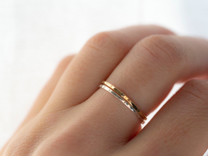 Puede incluir: Dos anillos finos de metal martillado, uno plateado y otro dorado, puestos en un dedo.