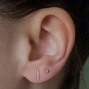 May include: Close-up of an ear with two gold earrings. One is a small, vertical bar, and the other is a tiny ball stud. The skin tone is light, and the earrings are simple and elegant.