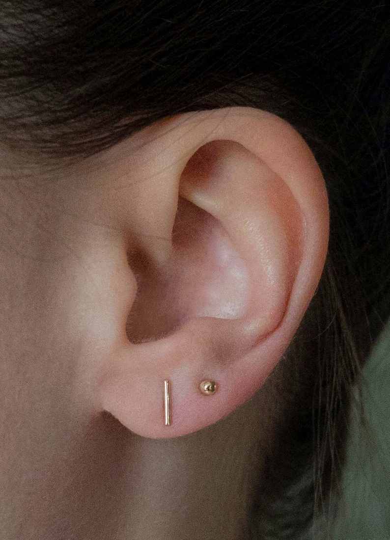 May include: Close-up of an ear with two gold earrings. One is a small, vertical bar, and the other is a tiny ball stud. The skin tone is light, and the earrings are simple and elegant.