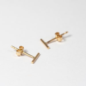 May include: A pair of gold-colored stud earrings. Each earring features a simple, straight bar design. The earrings are displayed on a white surface, with a focus on their minimalist aesthetic. The earrings are small and delicate.