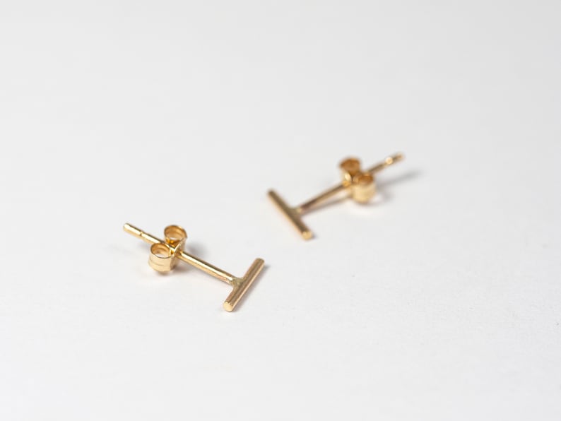 May include: A pair of gold-colored stud earrings. Each earring features a simple, straight bar design. The earrings are displayed on a white surface, with a focus on their minimalist aesthetic. The earrings are small and delicate.