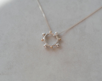 May include: A silver necklace with a circular pendant featuring a textured silver ring with gold accents. The pendant has a series of small, round beads around the edge.