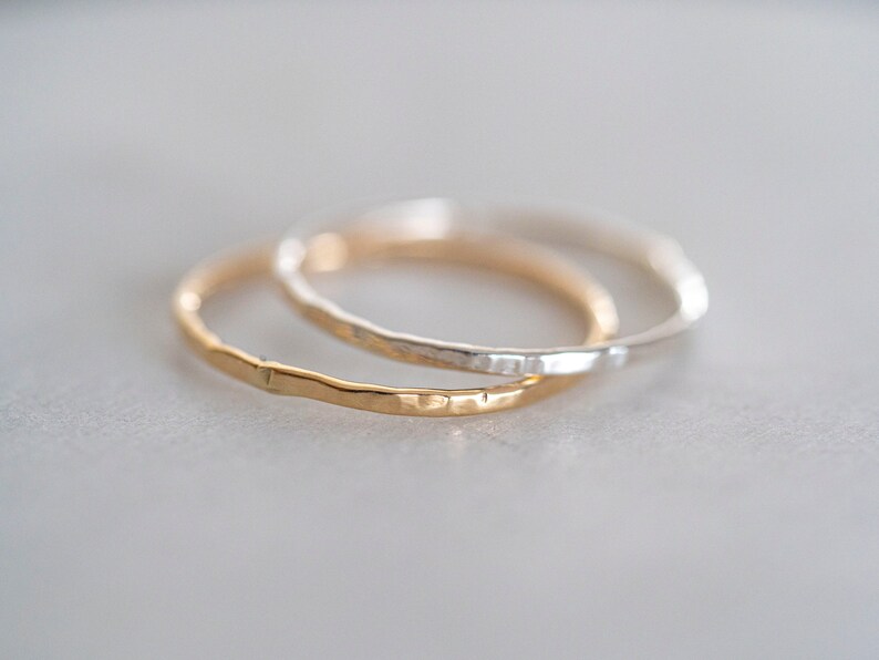 Puede incluir: Dos anillos finos de metal martillado, uno dorado y otro plateado, est&aacute;n apilados uno encima del otro.