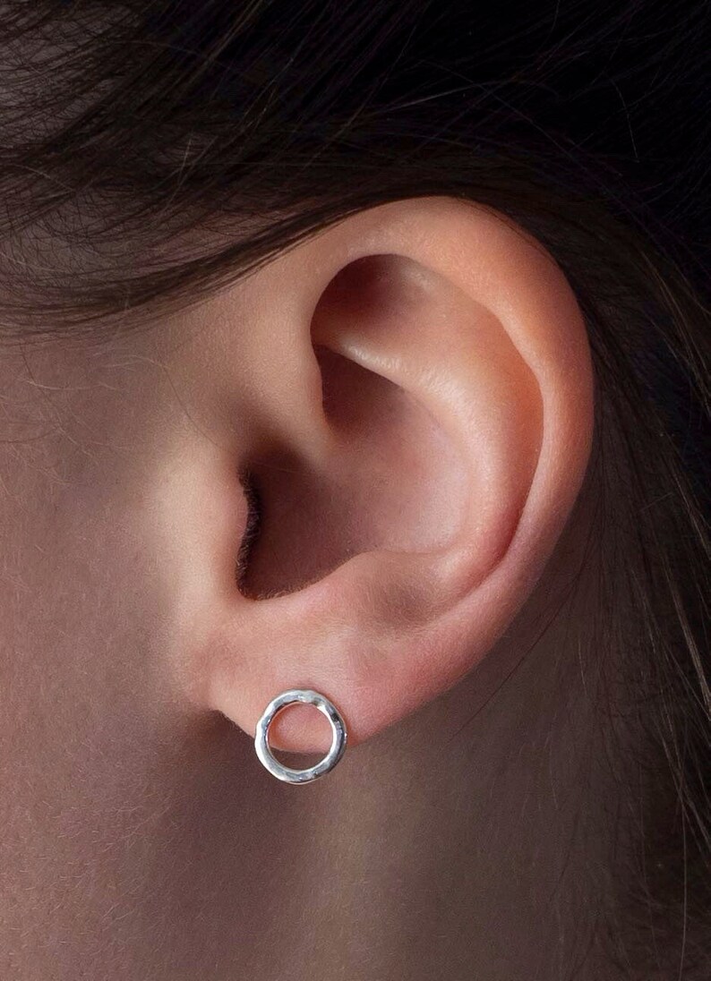 May include: A close-up of a person's ear with a small, silver, hammered circle earring.