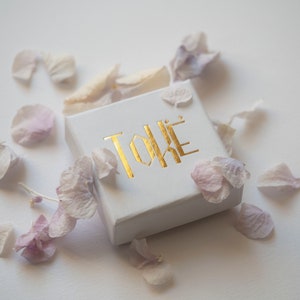 May include: A white box with the word "TOKE" printed in gold letters. The box is surrounded by pink and white flower petals.