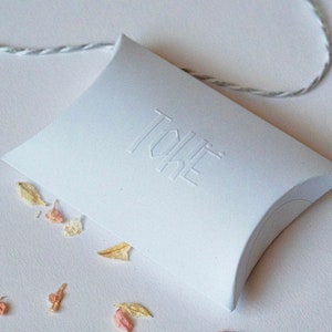 May include: White pillow box with the word "TOKE" embossed on the top. The box is sitting on a white surface with dried flower petals scattered around it.