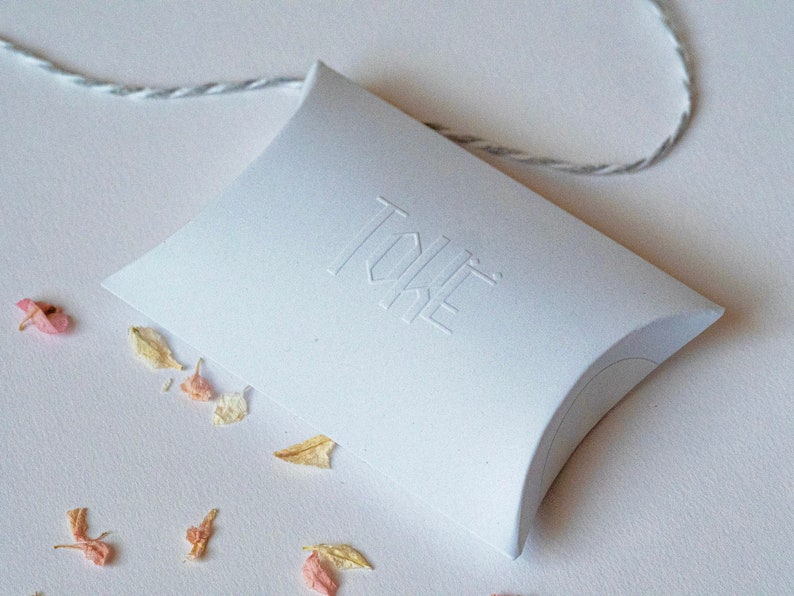 May include: White pillow box with the word "TOKE" embossed on the top. The box is sitting on a white surface with dried flower petals scattered around it.