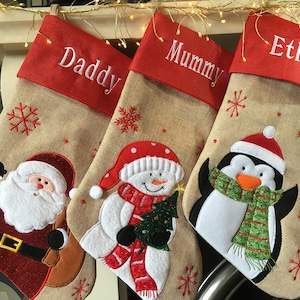 May include: Three personalised Christmas stockings with red felt tops and hessian bodies. The stockings feature embroidered names "Daddy", "Mummy", and "Ethan". Each stocking has a different festive applique: Father Christmas, a snowman, and a penguin.