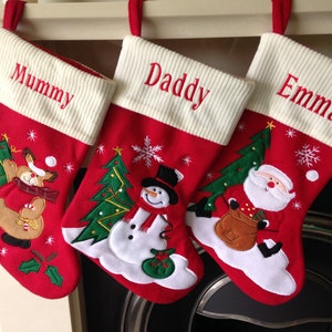 May include: Three red Christmas stockings with cream ribbed tops and red hanging loops. Each stocking is embroidered with a name: Mummy, Daddy, and Emma. Festive appliqués include a reindeer, snowman, and Santa Claus.